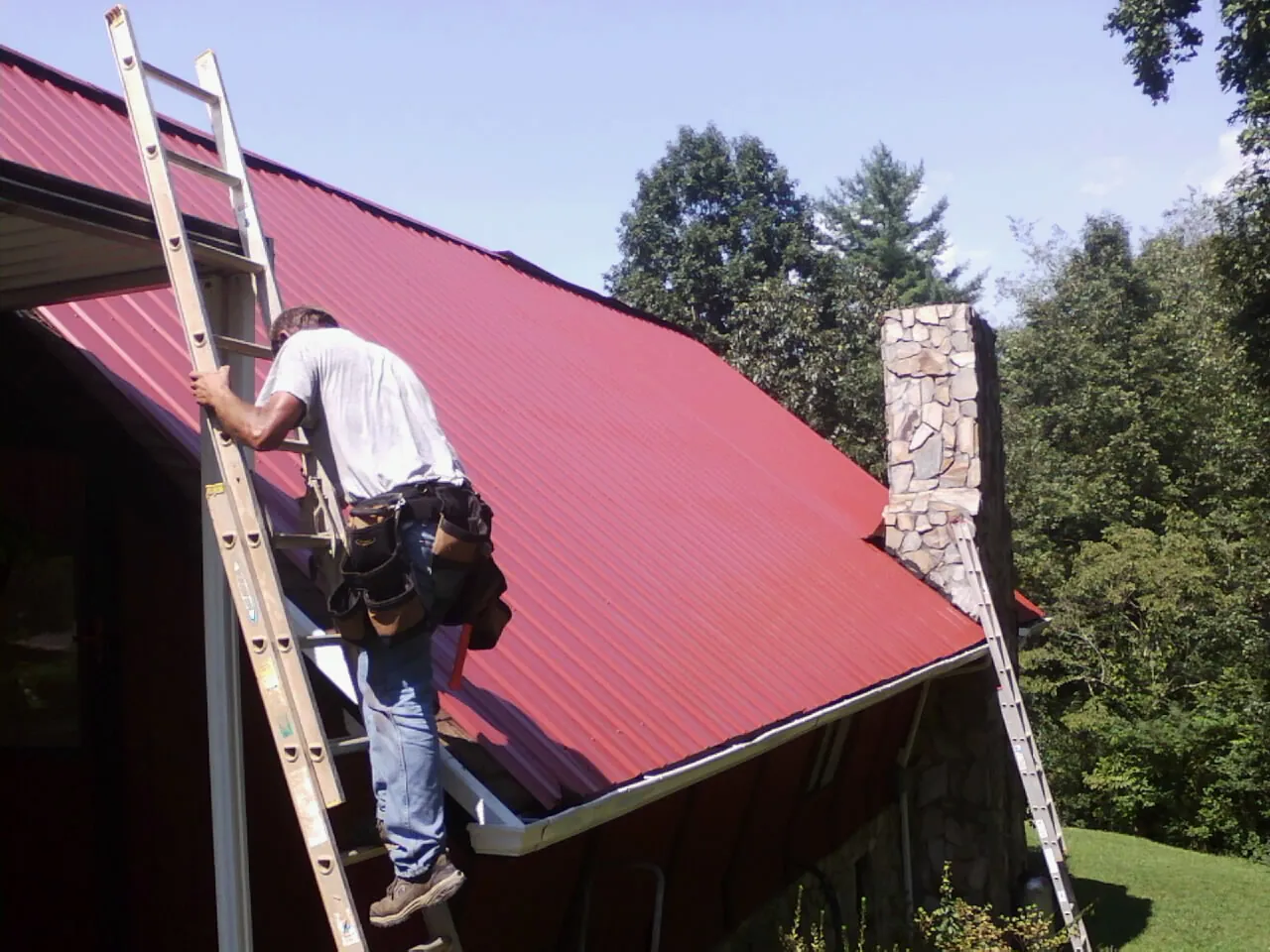 Roofer on a ladder beside a red metal roof and stone chimney — Soffit & Fascia Repair by Roofing Brothers in Eastham